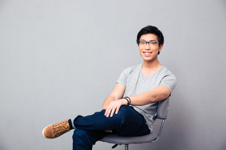 Smiling asian man sitting on the chair over gray background and looking at cameraの写真素材