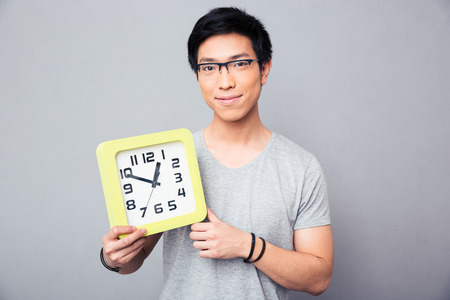 Happy asian man holding big clock over gray background and looking at camera