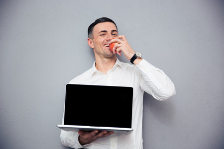 Happy businessman showing blank laptop screen and eating apple over gray background. Looking at...