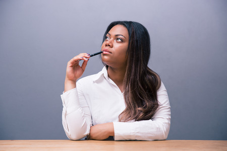 Pensive african businesswoman sitting at the table with pen and looking up over gray backgroundの写真素材