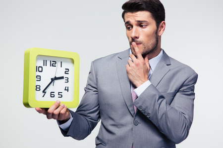 Pensive businessman holding clock over gray background