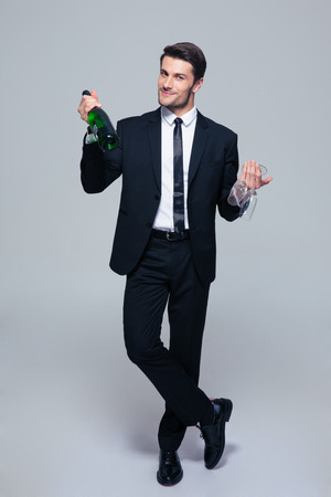 Full length portrait of a smiling businessman holding bottle with champagne and glass over gray backgroundの写真素材