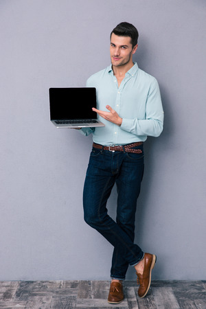 Full length portrait of a casual man showing blank laptop screen over gray backgroundの写真素材