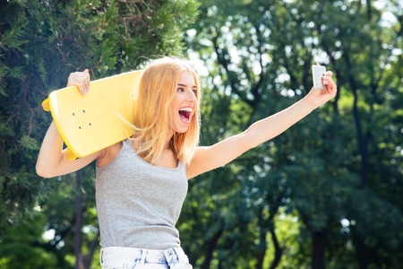 Funny young girl with skateboard making selfie photo on smartphone outdoorsの写真素材