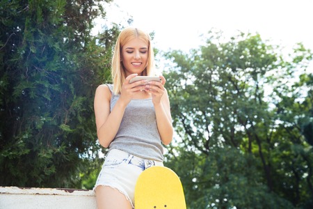 Happy young girl with skateboard using smartphone outdoorsの写真素材