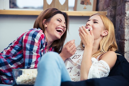Two cheerful girls lying on the sofa and eating popcorn at homeの写真素材