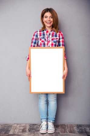 Full length portrait of a smiling casual woman holding blank board over gray backgroundの写真素材