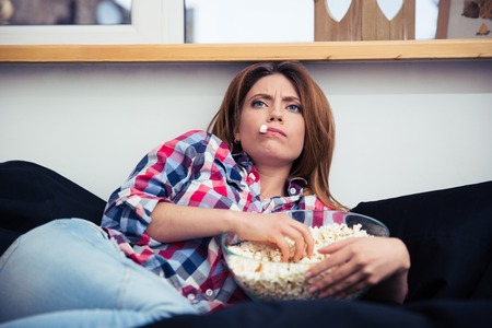 Young woman lying on the sofa and eating popcorn at homeの写真素材