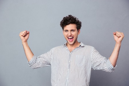 Successful young man celebrating his winning over gray background. Looking at camera