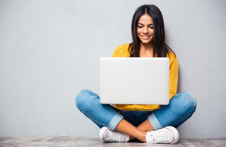 Happy young woman sitting on the floor with corssed legs and using laptop on gray background