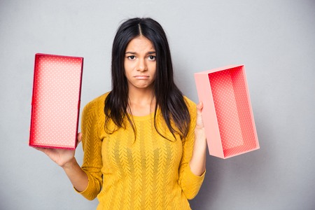 Unhappy woman holding empty gift box over gray background. Looking at camera