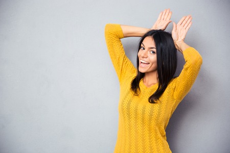 Happy woman showing rabbit ears with her palms over gray background. Looking at camera