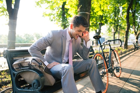 Smiling businessman sitting on the bench and talking on the phone in park outdoorsの写真素材