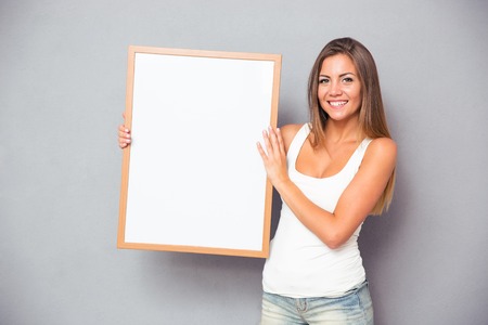 Happy casual woman holding blank board over gray background. Looking at cameraの写真素材