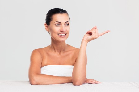 Happy beautiful woman in towel sitting at the table and pointing finger away isolated on a white backgroundの写真素材