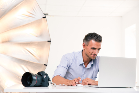 Portrait of a happy photographer using laptop at his workplaceの写真素材