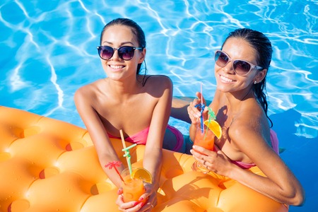 Portrait of a two smiling girlfriends drinking cocktails in swimming pool
