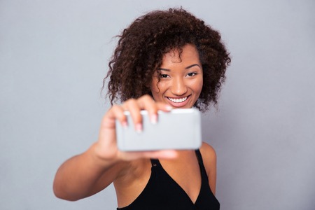 Portrait of a happy afro american woman making selfie photo on smartphone over gray backgroundの写真素材