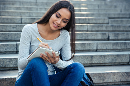 Portrait of a smiling female student sitting on the city stairs and writing notes in notepad outdoorsの写真素材