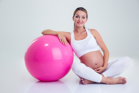Portrait of a smiling pregnant woman sitting on the floor with fitness ball isolated on a white backgroundの写真素材