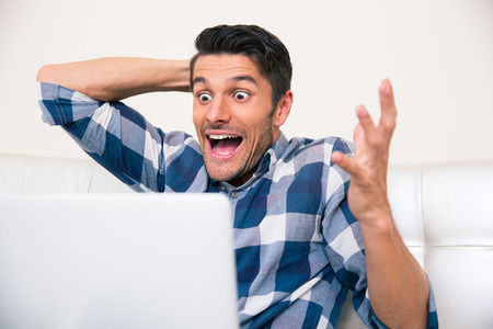 Portrait of a very excited man sitting on the sofa with laptop at homeの写真素材