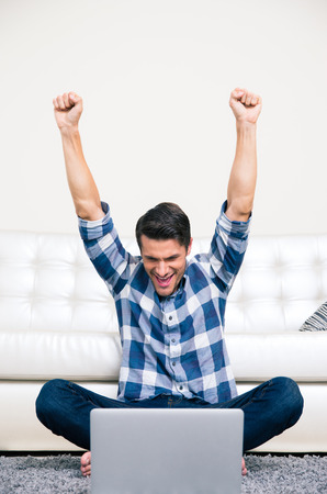 Portrait of excited man sitting on the floor and watching the game on laptop at homeの写真素材
