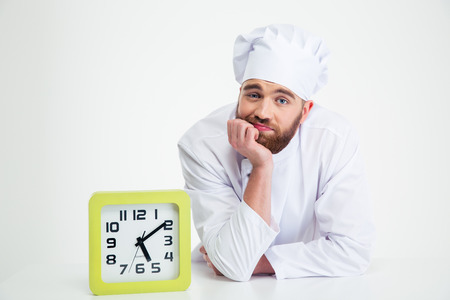 Portrait of a pensive chef cook leaning on the table with clock isolated on a white background
