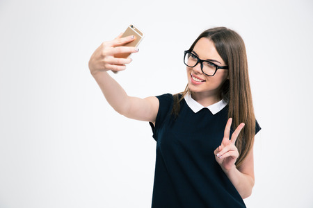 Portrait of a happy young girl making selfie photo on smartphone while showing victory sign isolated on a white backgroundの写真素材