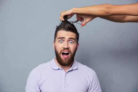 Portrait of a man looking scared while female hands cutting his hair over gray backgroundの写真素材