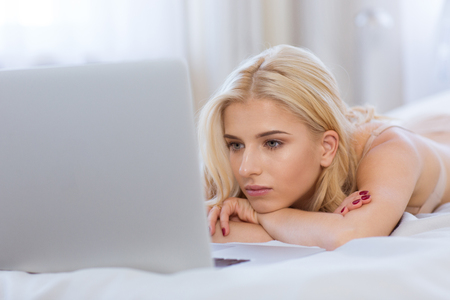 Portrait of a beautiful blonde woman using laptop computer on the bed at homeの写真素材