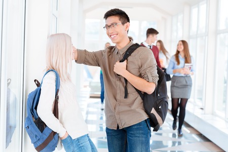 Asian boy in black glasses and blond girl are carrying backpacks and talking in the corridorの写真素材