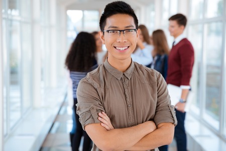 Portrait of a happy male student standing with arms folded in university hall with classmated on...