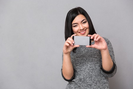 Beautiful happy smiling asian girl in gray jumper taking photos using cellphoneの写真素材