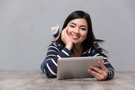 Portrait of a smiling woman lying on the floor with tablet computer on gray background and looking at cameraの写真素材