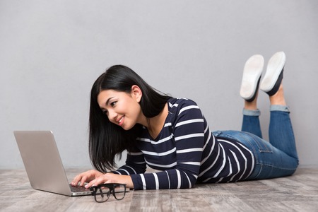 Portrait of a happy beautiful woman lying on the floor and using laptop computer on gray backgroundの写真素材