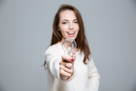 Portrait of a happy woman holding bulb over gray background. Focus on bulbの写真素材