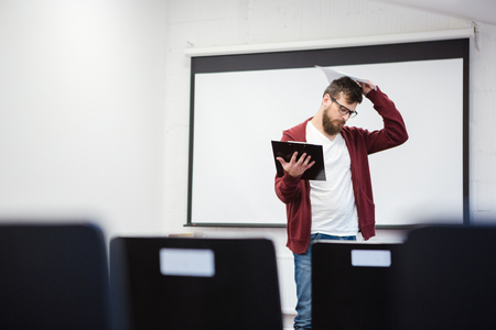 Young teacher with beard in brown hoodie thinking in empty classroom near the whiteboardの写真素材