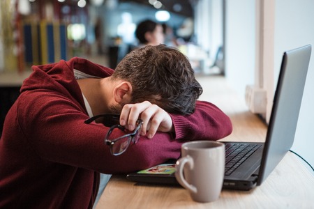 Tired man in brown sweetshirt holding glasses while sleeping at office deskの写真素材