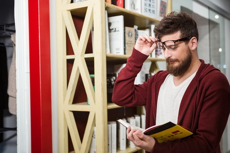 Bearded  young handsome man in brown hoodie and glasses reading book standing near the wooden bookshelfの写真素材