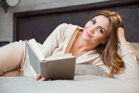 Beautiful young woman in pajamas relaxing on the bed and reading bookの写真素材