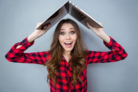 Funny pretty smiling girl in plaid shirt holding laptop above her headの写真素材