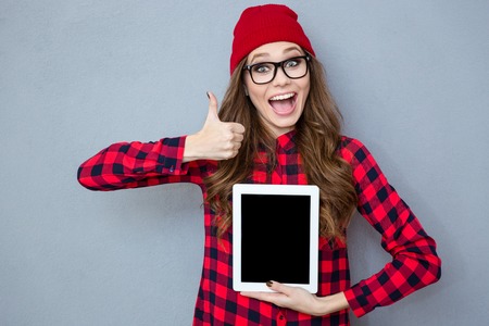Portrait of a cheerful woman showing blank tablet computer screen and thumb up on gray backgroundの写真素材