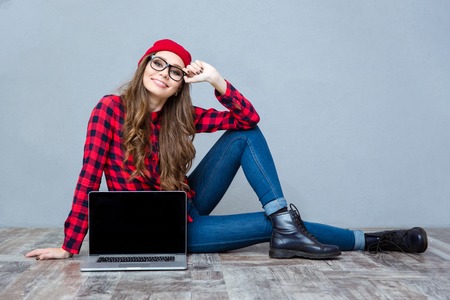 Portrait of a happy casual woman sitting on the floor and showing blank laptop computer screen on gray backgroundの写真素材