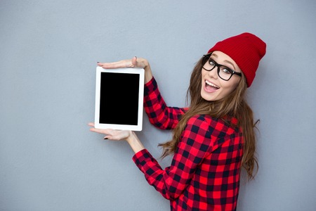 Portrait of a happy hipster woman showing blank tablet computer screen on gray backgroundの写真素材
