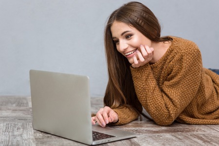 Pretty attractive sweet cheerful young woman with long hair in brown sweater lying on wooden floor using laptopの写真素材