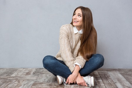 Happy excited young woman with long hair in beige sweater and jeans sitting on wooden floor with legs crossed and looking awayの写真素材