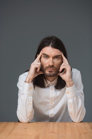 Pensive man with beard and long hair sitting at the table and looking at camera over gray backgroundの写真素材