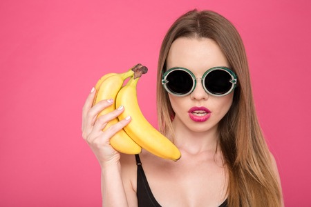 Closeup of beautiful attractive concenrated young female in sunglasses and black swimsuit holding bananas on pink backgroundの写真素材