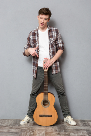 Full length portrait of a young man winking and pointing finger on guitar on gray backgroundの写真素材