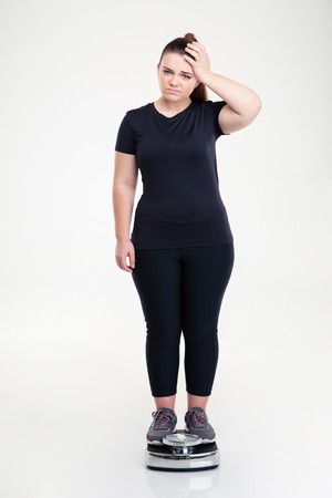 Full length portrait of a sad thick woman standing on weighing machine isolated on a white backgroundの写真素材